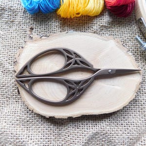 May include: Decorative embroidery scissors with a detailed floral design, resting on a wooden slice. The scissors are silver-toned and feature intricate cut-out patterns. Colorful embroidery floss and a wooden embroidery hoop are in the background.