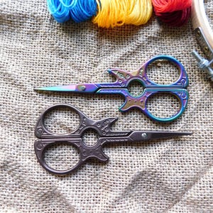 May include: Two pairs of decorative scissors with intricate designs. One pair is iridescent with a guitar-shaped handle, the other is brown with a more traditional design. The scissors are on a burlap background.