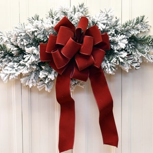 May include: A Christmas swag with snow-dusted fir branches and a large, deep red velvet bow. The bow has multiple loops and long tails, creating a classic festive decoration for the holiday season.