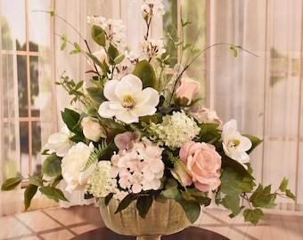 May include: A gold metal vase with a round top and a pedestal base holds a floral arrangement of pink and white roses, white hydrangeas, white magnolia blossoms, and white cherry blossoms.