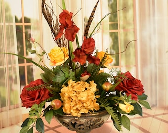 May include: A floral arrangement in a gold-toned, ornate vase. The arrangement features red roses, yellow hydrangeas, yellow roses, and red irises. The arrangement is accented with green leaves and brown feathers.