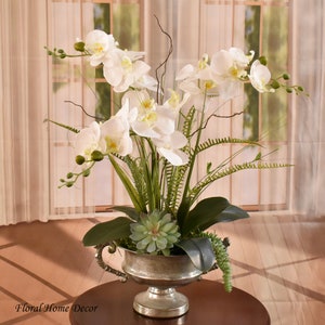 Könnte beinhalten: Weiße Orchideen-Blumenarrangement in einer silbernen Vase mit grünen Sukkulenten. Das Arrangement steht auf einem Holztisch.