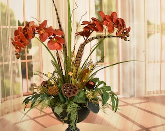 May include: A red orchid flower arrangement in a dark brown urn vase. The arrangement includes green foliage, brown feathers, and dried seed pods.
