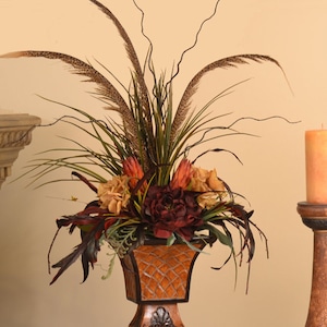 May include: A brown and gold ceramic vase with a square base and a decorative design. The vase is filled with a floral arrangement of red, orange, and brown flowers, as well as green leaves and brown feathers.