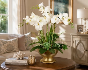May include: An artificial floral arrangement featuring white orchid blossoms with green buds, green foliage, and a small succulent, all contained within a ribbed, metallic gold-toned pedestal bowl with a wide, circular base and flared rim.