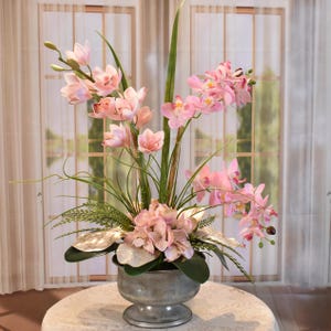 以下が含まれることがあります： ピンクの蘭やその他の花をあしらった、装飾的な古びた金属の花瓶。緑の葉と細長い葉が自然なディスプレイを作り出しています。花瓶は白い表面に置かれています。
