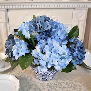 May include: A floral arrangement featuring blue hydrangea flowers in a decorative blue and white ceramic vase. The arrangement includes green leaves and is set on a table with a white tablecloth.