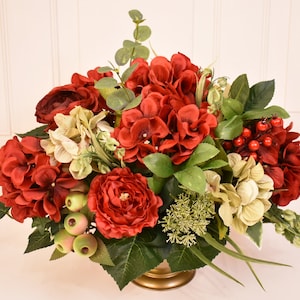 May include: A red and green floral arrangement in a gold vase. The arrangement features red hydrangeas, red roses, and green foliage.