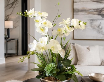 May include: A decorative arrangement featuring white orchid flowers with yellow centers, green succulent plants, and dark green leaves. The botanical display is contained within a rounded, metallic silver bowl with a smooth, reflective finish.