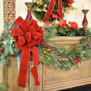 May include: A red velvet bow tied on a green garland with pine needles, red berries, and pine cones. The garland is draped over a wooden mantel.