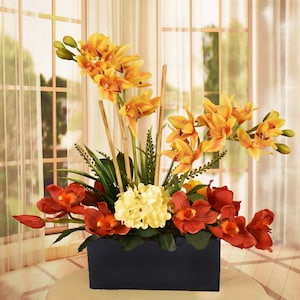 May include: A floral arrangement featuring vibrant orange and red orchids, accented by bamboo stalks and greenery. The flowers are arranged in a rectangular black container, creating a striking visual contrast. The arrangement is set against a window with sheer curtains.