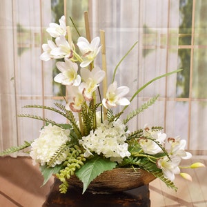 May include: A white orchid and hydrangea floral arrangement in a gold bowl. The arrangement features white flowers with green leaves and stems.