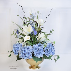 May include: A blue and white floral arrangement in a gold vase. The arrangement features hydrangeas, white magnolias, and other flowers. The vase has a round base and a flared top.