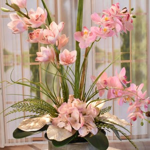 以下が含まれることがあります： 明るいピンク色の蘭と紫陽花を銀色の花瓶にアレンジしたフラワーアレンジメント。緑の葉と銀色の葉が含まれています。背景は、シアーなカーテンのある窓です。