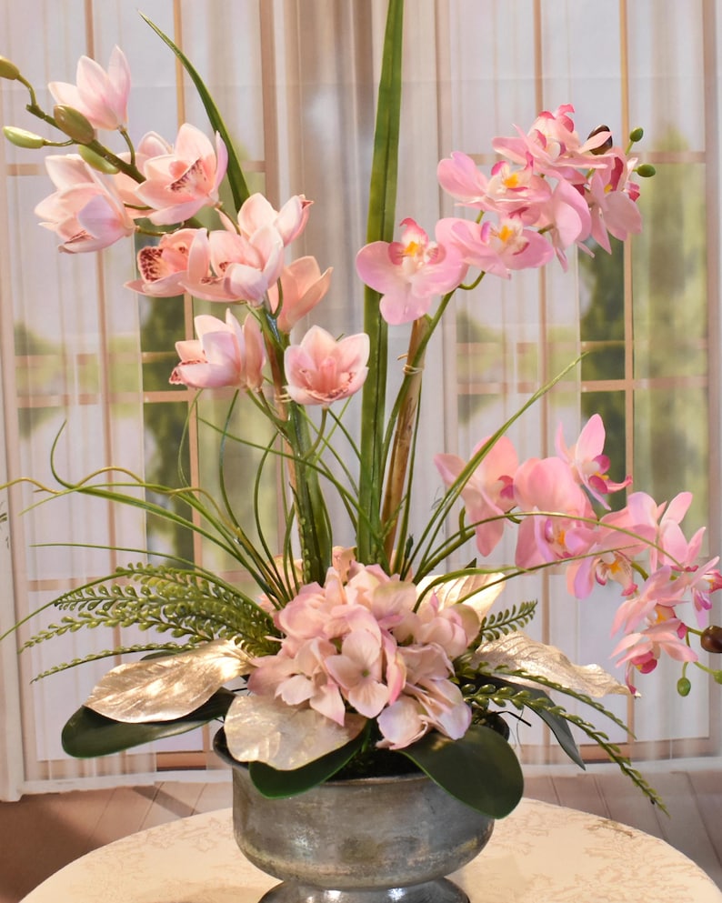 以下が含まれることがあります： 明るいピンク色の蘭と紫陽花を銀色の花瓶にアレンジしたフラワーアレンジメント。緑の葉と銀色の葉が含まれています。背景は、シアーなカーテンのある窓です。