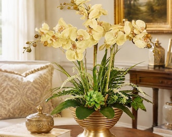 May include: A decorative arrangement of pale yellow artificial orchid flowers with green foliage and a small succulent plant, housed in a ribbed, metallic gold-toned pedestal bowl. The floral display features multiple stems and buds.