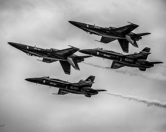 May include: Four black and white fighter jets fly in formation, leaving trails of smoke in the sky. The jets are labeled "Blue Angels" and "U.S. Navy".