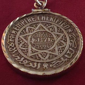 May include: A gold pendant necklace with a round coin-shaped charm featuring a six-pointed star with intricate details and the inscription "EMPIRE CHERIFIEN" and "1371" around the star.