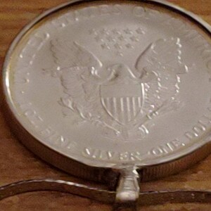 May include: A silver coin pendant featuring an eagle design. The coin is inscribed with the words "UNITED STATES OF AMERICA" and "ONE DOLLAR". The pendant is attached to a silver chain. The coin is approximately 1.5 inches in diameter.