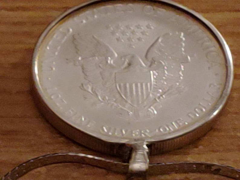 May include: A silver coin pendant featuring an eagle design. The coin is inscribed with the words "UNITED STATES OF AMERICA" and "ONE DOLLAR". The pendant is attached to a silver chain. The coin is approximately 1.5 inches in diameter.