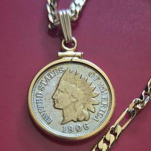 Puede incluir: Un collar de tono dorado con un colgante circular que contiene una moneda de un centavo de los Estados Unidos de 1906. La moneda muestra el perfil de una persona con un tocado, rodeado por el texto UNITED STATES OF AMERICA y 1906.