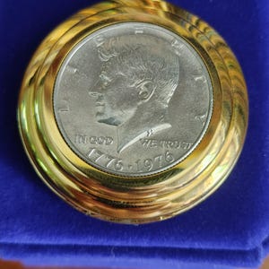 May include: A gold-toned pocket watch case featuring a silver-colored 1776-1976 Kennedy half-dollar coin as the face. The coin displays the profile of John F. Kennedy, the text LIBERTY, IN GOD WE TRUST, and the dual dates 1776-1976. A gold loop is attached.