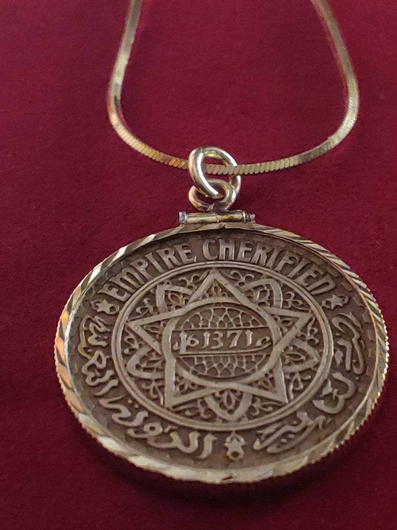 May include: A gold pendant necklace with a round coin-shaped charm featuring a six-pointed star with intricate details and the inscription "EMPIRE CHERIFIEN" and "1371" around the star.