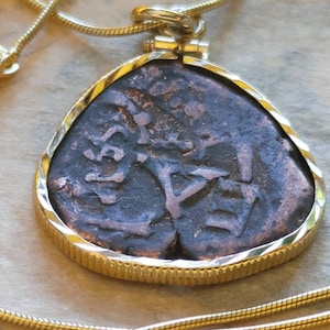 May include: A gold-coloured necklace with a pendant holding a dark, textured coin. The coin is set in a gold-coloured frame with a textured edge. The chain is a thin, gold-coloured snake chain, creating an elegant and historical aesthetic.