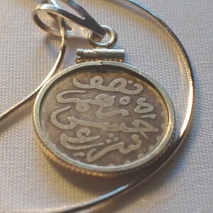 May include: A silver chain necklace with a round pendant featuring Arabic script. The pendant is made of a silver coin with a gold rim.