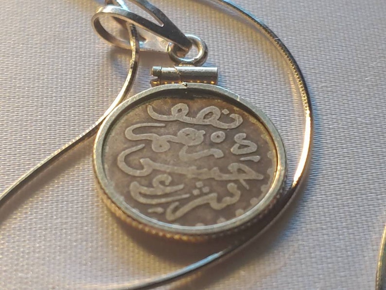 May include: A silver chain necklace with a round pendant featuring Arabic script. The pendant is made of a silver coin with a gold rim.