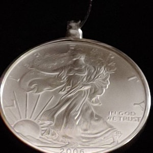 May include: A silver coin pendant with a detailed relief of a woman walking towards the sun, with the text "IN GOD WE TRUST" and the year "2006". The pendant is attached to a black string.