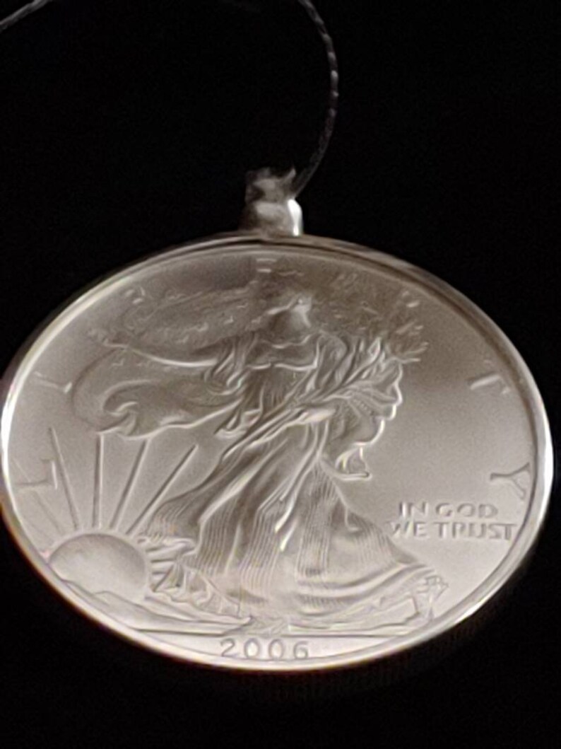 May include: A silver coin pendant with a detailed relief of a woman walking towards the sun, with the text "IN GOD WE TRUST" and the year "2006". The pendant is attached to a black string.