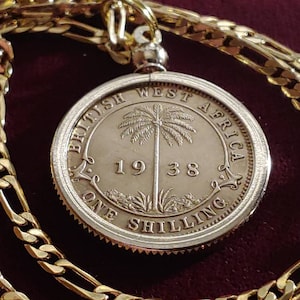 May include: A gold-colored chain necklace with a pendant featuring a 1938 British West Africa coin. The coin is silver-colored and has a palm tree design in the center. The chain has a textured, linked design.