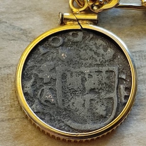 Puede incluir: Un colgante dorado con una moneda oscura y texturizada. La moneda está engastada en un marco dorado circular y suspendida de una cadena dorada. La superficie de la moneda muestra un diseño en relieve, posiblemente un escudo de armas o un emblema histórico.