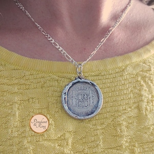 Op de afbeelding: Een zilveren ketting met een munt hanger. De munt, gezet in een decoratieve zilveren lijst, toont gedetailleerde afbeeldingen. De ketting is een schakelontwerp. De persoon draagt een gele trui.