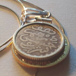 May include: A gold chain necklace with a pendant featuring an old coin with Arabic script. The coin is encased in a gold frame.