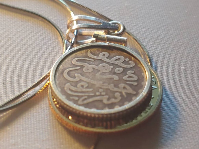 May include: A gold chain necklace with a pendant featuring an old coin with Arabic script. The coin is encased in a gold frame.