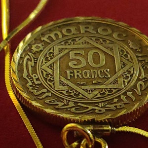May include: A gold coin with the inscription "MAROC" and "50 Francs" in the centre. The coin is surrounded by an intricate design and is attached to a gold chain.