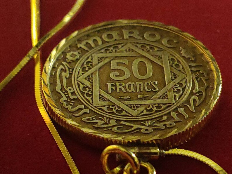 May include: A gold coin with the inscription "MAROC" and "50 Francs" in the centre. The coin is surrounded by an intricate design and is attached to a gold chain.
