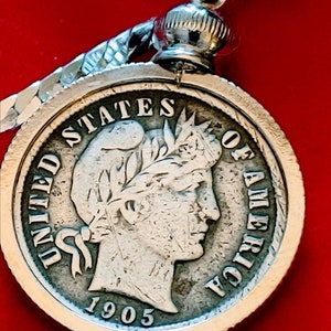 May include: A silver coin pendant featuring a detailed profile of a woman with a laurel wreath, inscribed with "UNITED STATES OF AMERICA" and the year "1905". The pendant is attached to a silver chain, set against a red background.