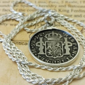 Op de afbeelding: Een zilverkleurige ketting met een ronde hanger. De hanger heeft een reliëfontwerp, inclusief een wapen en tekst. De ketting is een strak geweven touwstijl. De achtergrond is een vintage papier.