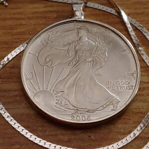 May include: A silver coin pendant necklace featuring a 2006 American Silver Eagle coin. The coin depicts Lady Liberty and the sun, with the inscription "IN GOD WE TRUST." The necklace has a delicate silver chain.