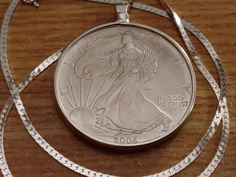May include: A silver coin pendant necklace featuring a 2006 American Silver Eagle coin. The coin depicts Lady Liberty and the sun, with the inscription "IN GOD WE TRUST." The necklace has a delicate silver chain.