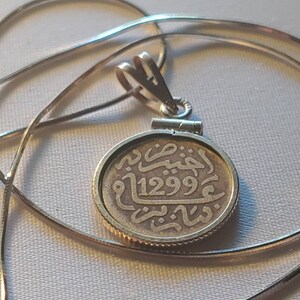 May include: Silver pendant necklace with a round coin charm featuring Arabic script and the date 1299. The coin is attached to a silver chain.