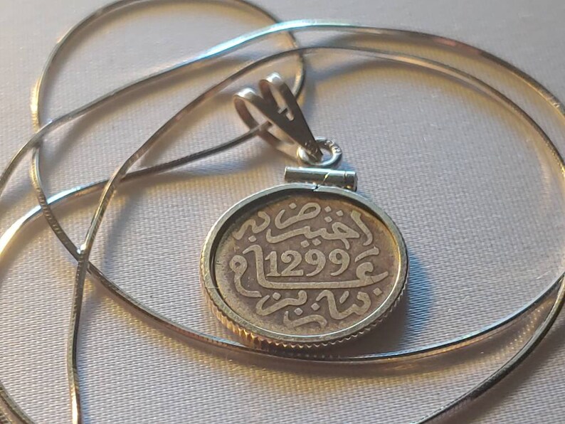 May include: Silver pendant necklace with a round coin charm featuring Arabic script and the date 1299. The coin is attached to a silver chain.