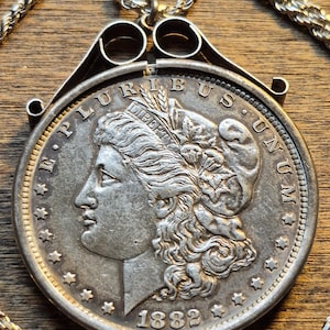 Peut inclure: Pendentif pièce de monnaie en argent avec un profil détaillé de femme, l'année 1882 et l'inscription "E PLURIBUS UNUM". La pièce est sertie dans un cadre argenté décoratif et suspendue à une chaîne argentée. Le pendentif est présenté sur une surface en bois.