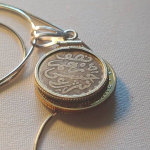 May include: A silver necklace with a pendant featuring a gold-rimmed coin with Arabic script. The coin is attached to the necklace with a silver clasp.