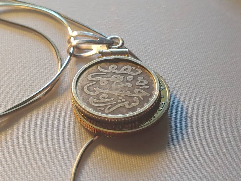 May include: A silver necklace with a pendant featuring a gold-rimmed coin with Arabic script. The coin is attached to the necklace with a silver clasp.