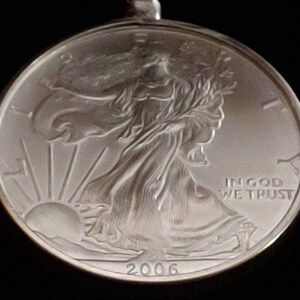May include: A silver coin pendant featuring the Walking Liberty design. The coin is dated 2006 and includes the inscription "IN GOD WE TRUST". The pendant is round and has a loop for a chain.