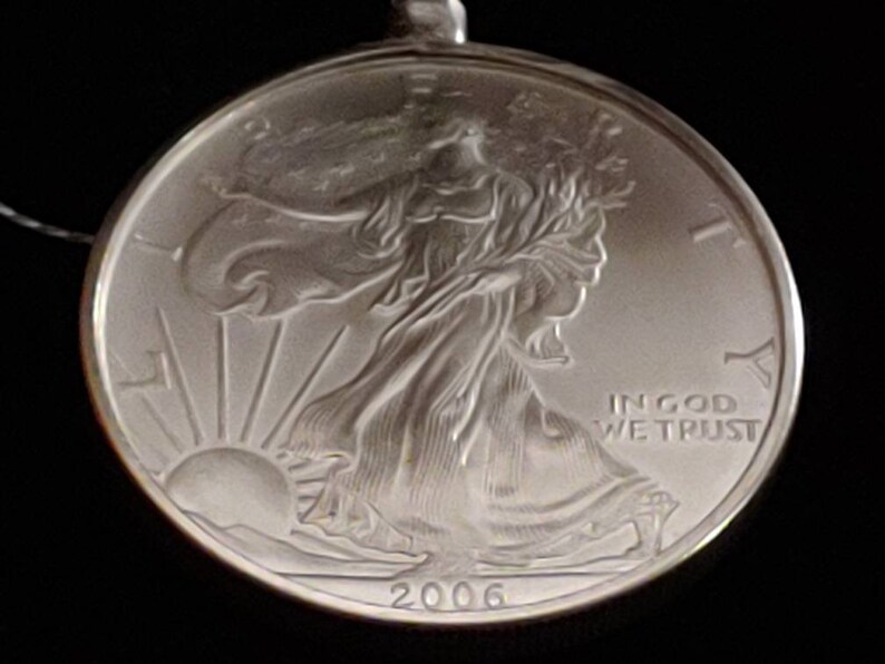 May include: A silver coin pendant featuring the Walking Liberty design. The coin is dated 2006 and includes the inscription "IN GOD WE TRUST". The pendant is round and has a loop for a chain.